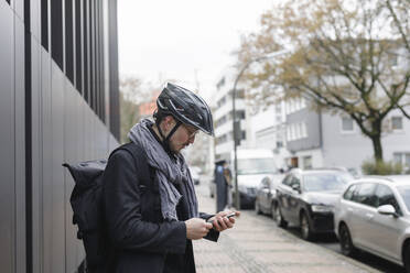 Junger Mann mit Fahrradhelm und Rucksack steht auf dem Bürgersteig und schaut auf sein Handy - KMKF01154