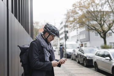 Young man with cycling helmet and backpack standing on pavement looking at cell phone - KMKF01154