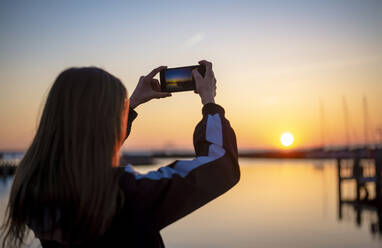 Deutschland, Mecklenburg-Vorpommern, Insel Poel, Rückansicht eines jugendlichen Mädchens beim Fotografieren des Meeres bei Sonnenuntergang - BFRF02170