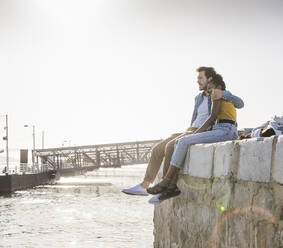 Young couple sitting on pier at the waterfront enjoying the view, Lisbon, Portugal - UUF19811