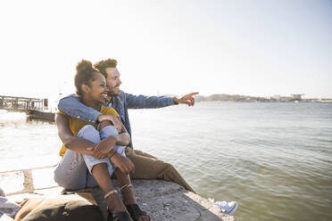 Glückliches junges Paar auf dem Pier am Wasser sitzend mit Blick auf die Aussicht, Lissabon, Portugal - UUF19816