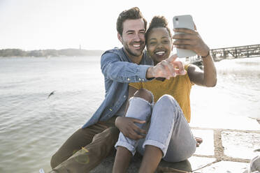 Happy young couple sitting on pier at the waterfront taking a selfie, Lisbon, Portugal - UUF19818