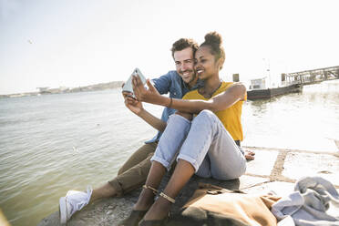Glückliches junges Paar sitzt auf dem Pier am Wasser und macht ein Selfie, Lissabon, Portugal - UUF19820