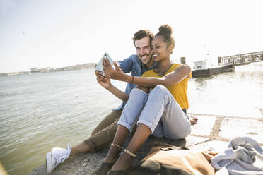 Happy young couple sitting on pier at the waterfront taking a selfie, Lisbon, Portugal - UUF19820