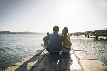 Rückansicht eines jungen Paares, das auf einem Pier am Wasser sitzt, Lissabon, Portugal - UUF19821