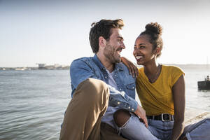 Happy young couple sitting on pier at the waterfront, Lisbon, Portugal - UUF19834