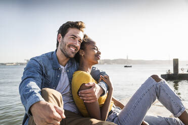 Happy young couple sitting on pier at the waterfront, Lisbon, Portugal - UUF19836