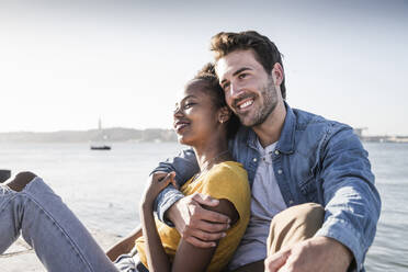 Happy young couple sitting on pier at the waterfront, Lisbon, Portugal - UUF19837