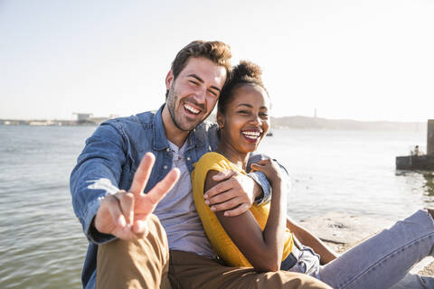Porträt eines glücklichen jungen Paares, das am Ufer sitzt und gestikuliert, Lissabon, Portugal, lizenzfreies Stockfoto