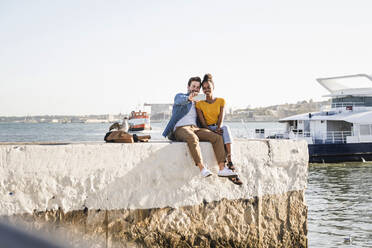 Glückliches junges Paar sitzt auf dem Pier am Wasser und macht ein Selfie, Lissabon, Portugal - UUF19842
