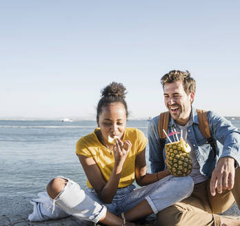 Glückliches junges Paar, das mit einer Ananas auf dem Pier am Wasser sitzt, Lissabon, Portugal - UUF19850