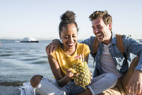 Happy young couple sitting on pier at the waterfront with a pineapple, Lisbon, Portugal - UUF19851