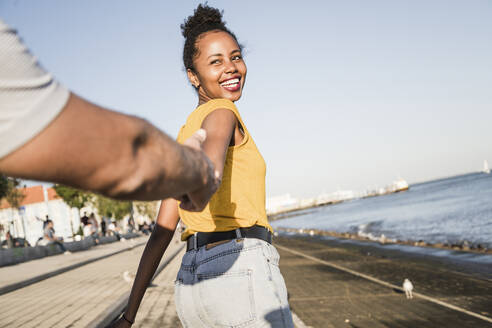 Happy young woman holding hand of her boyfriend on pier at the waterfront, Lisbon, Portugal - UUF19861