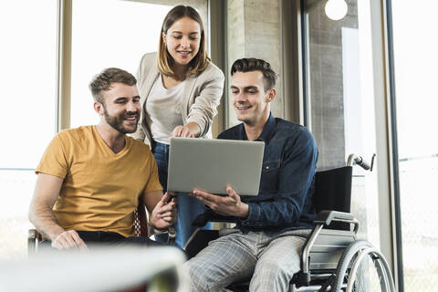 Junger Geschäftsmann im Rollstuhl zeigt Kollegen im Büro seinen Laptop, lizenzfreies Stockfoto