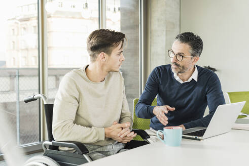 Mature businessman and young man in wheelchair talking at desk in office - UUF19948