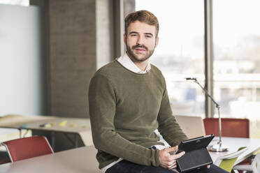 Portrait of a confident young businessman sitting on conference table in office - UUF19952