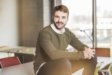 Portrait of a confident young businessman sitting on conference table in office - UUF19954