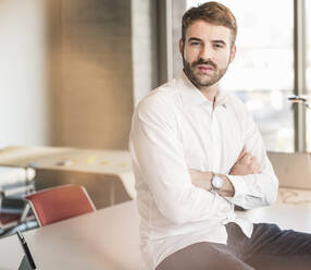 Portrait of a confident young businessman sitting on conference table in office - UUF19955