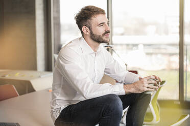 Young businessman sitting on conference table in office holding cup of coffee - UUF19957