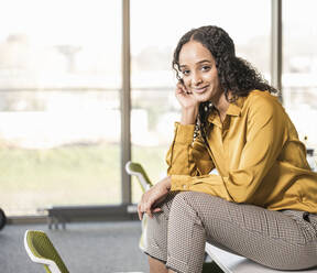 Portrait of smiling young businesswoman sitting on desk in office - UUF19959