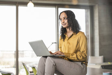 Young businesswoman sitting on desk in office using laptop - UUF19962