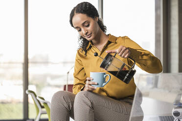 Junge Geschäftsfrau sitzt auf einem Schreibtisch im Büro und macht eine Kaffeepause - UUF19966