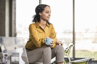 Young businesswoman sitting on desk in office having a coffee break - UUF19967