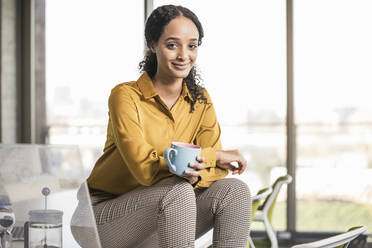 Portrait of smiling young businesswoman sitting on desk in office having a coffee break - UUF19968
