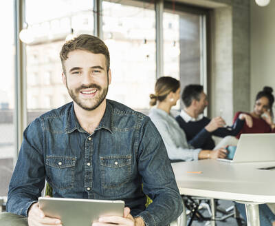Porträt eines lächelnden jungen Geschäftsmannes mit Tablet während einer Besprechung im Büro - UUF20018
