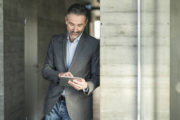 Mature businessman leaning against a wall in office using tablet - UUF20042