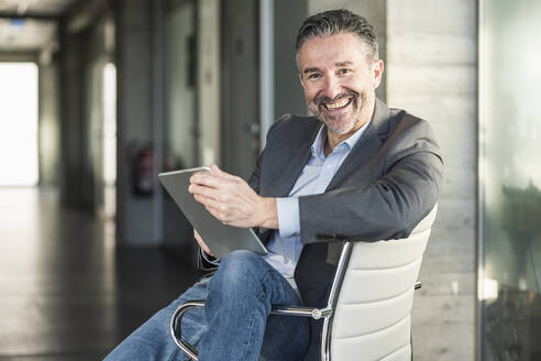 Porträt eines glücklichen reifen Geschäftsmannes mit Tablet sitzt auf Stuhl im Büro - UUF20045