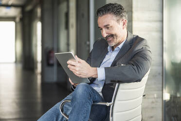 Mature businessman sitting on chair in office using tablet - UUF20046