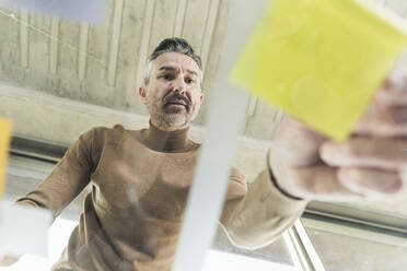 Mature businessman working on adhesive notes on glass table in office - UUF20050