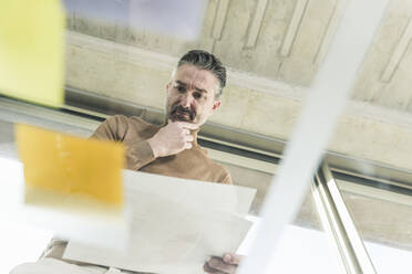 Mature businessman working on adhesive notes on glass table in office - UUF20051