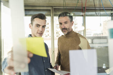 Reifer Geschäftsmann und junger Mann arbeiten an Haftnotizen an einer Glasscheibe im Büro - UUF20052