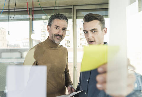 Reifer Geschäftsmann und junger Mann arbeiten an Haftnotizen an einer Glasscheibe im Büro, lizenzfreies Stockfoto