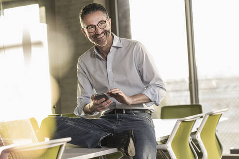 Glücklicher reifer Geschäftsmann mit Mobiltelefon im Büro - UUF20058