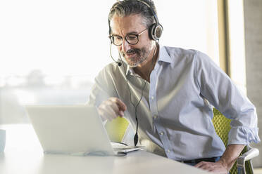 Mature businessman wearing headphones using laptop at desk in office - UUF20066