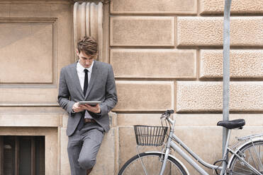 Junger Geschäftsmann lehnt an der Fassade und schaut auf ein digitales Tablet - FMOF00851