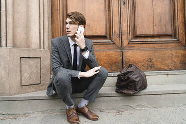 Portrait of young businessman on the phone sitting on steps of house entrance - FMOF00854