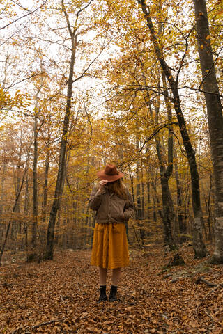 Modische rothaarige junge Frau im herbstlichen Wald, die ihr Gesicht mit einem Hut bedeckt, lizenzfreies Stockfoto