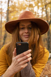 Porträt einer lächelnden rothaarigen jungen Frau im herbstlichen Wald, die auf ihr Handy schaut - AFVF04895