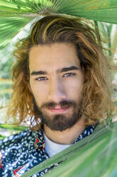 Porträt eines bärtigen jungen Mannes mit gefärbtem Haar zwischen Palmenblättern - AFVF04903