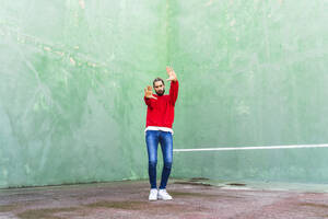 Portrait of serious young man wearing red sweatshirt raising hands in front of green wall - AFVF04925