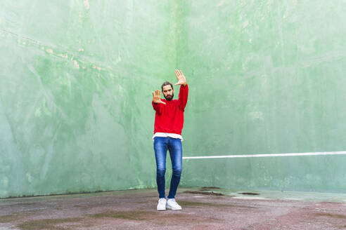 Porträt eines ernsten jungen Mannes in rotem Sweatshirt, der vor einer grünen Wand die Hände hebt - AFVF04925