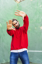 Portrait of serious young man wearing red sweatshirt raising hands in front of green wall - AFVF04926