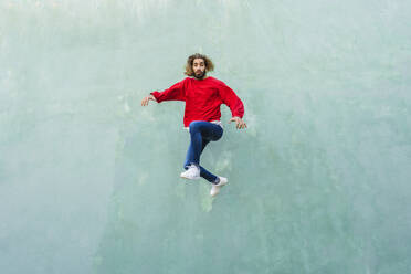 Portrait of young man wearing red sweatshirt jumping in the air in front of green wall - AFVF04938