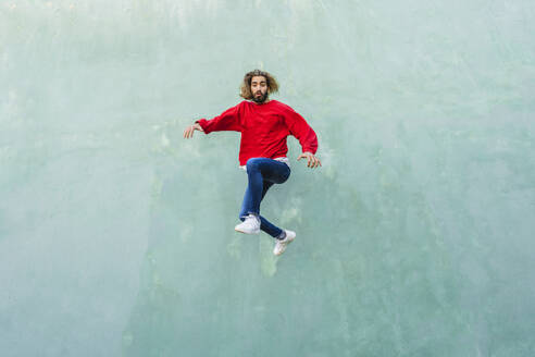 Porträt eines jungen Mannes mit rotem Sweatshirt, der vor einer grünen Wand in die Luft springt - AFVF04938