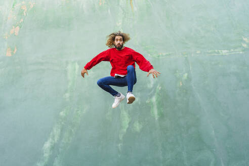 Porträt eines jungen Mannes mit rotem Sweatshirt, der vor einer grünen Wand in die Luft springt - AFVF04939