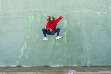 Porträt eines jungen Mannes in einem roten Sweatshirt, der vor einer grünen Wand in die Luft springt - AFVF04940
